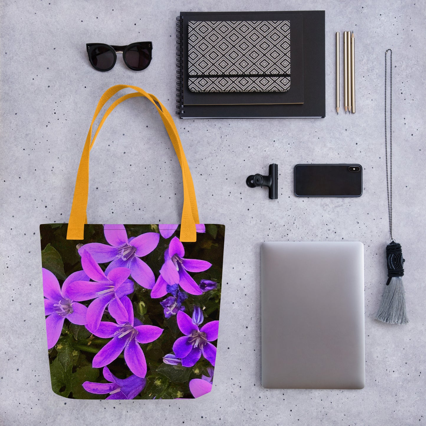 Eco-friendly floral bag with purple flowers and yellow handles displayed with sunglasses and tech accessories