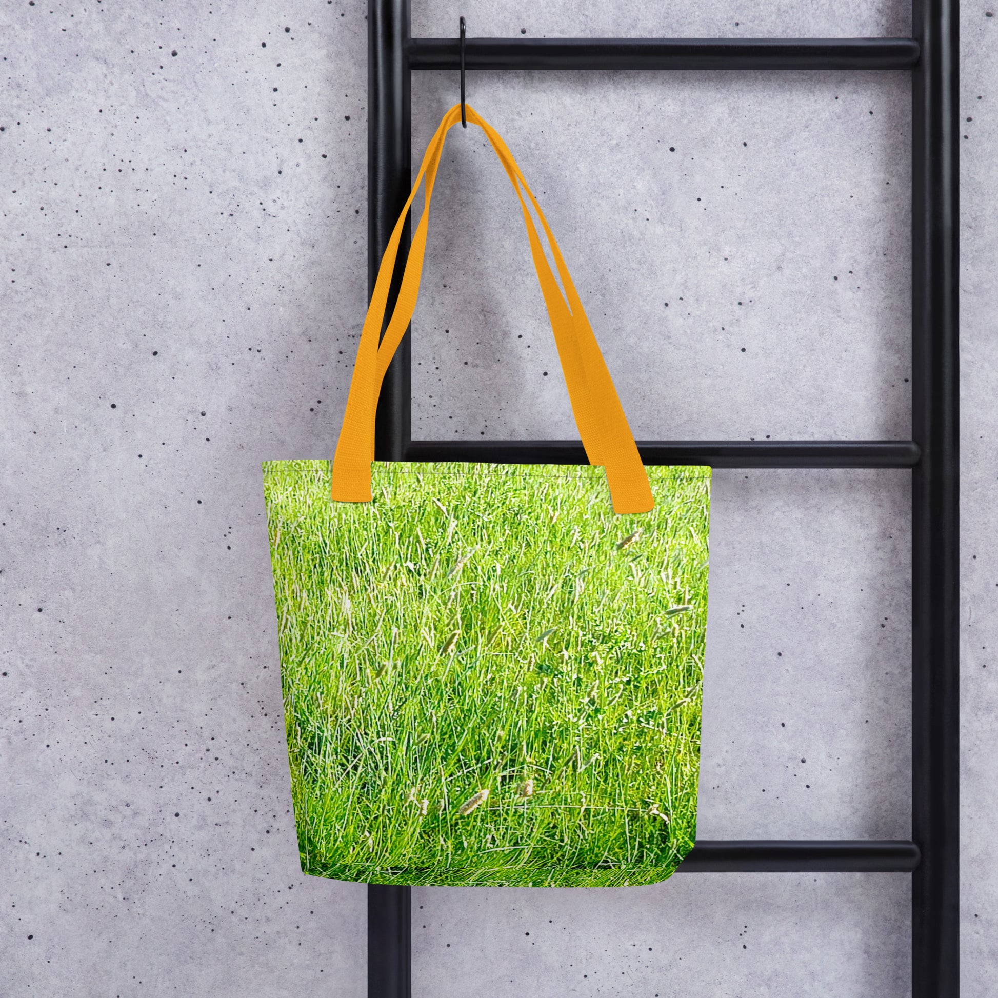 Grass print tote bag with bright yellow handles hanging on black metal ladder against gray wall